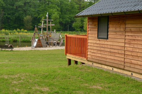 Wooden lodge with a playground by a river at Camping de Koeksebelt, Netherlands, surrounded by greenery.