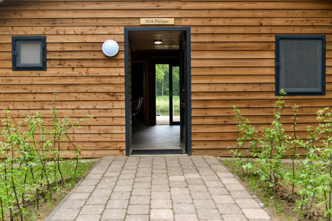 Entrada a River Lodge en Camping de Koeksebelt, Países Bajos, con fachada de madera y sendero de piedra.