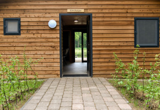 Entrada a River Lodge en Camping de Koeksebelt, Países Bajos, con fachada de madera y sendero de piedra.