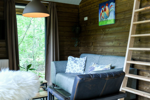 Salon confortable dans la River Lodge au Camping de Koeksebelt, avec canapé, échelle et vue sur la forêt.