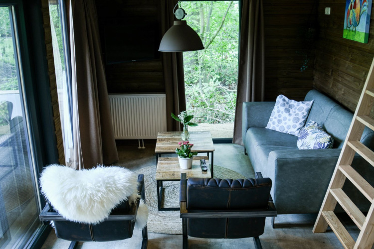 Gemütlicher Wohnraum mit großen Fenstern, Sofa und Sesseln in der River Lodge bei Camping de Koeksebelt.