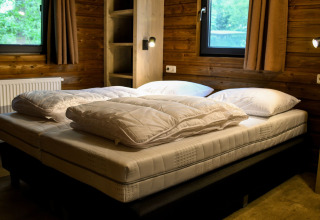 Soveværelse med dobbeltseng, vinduer med gardiner og træpaneler i River Lodge på Camping de Koeksebelt.