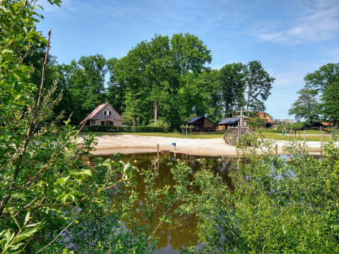 River Lodge at Camping de Koeksebelt in the Netherlands with sandy beach, lake, cabins and lush green trees.