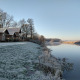 Frost-covered grass and wooden lodges by the river at River Cottage, Camping de Koeksebelt, Netherlands.