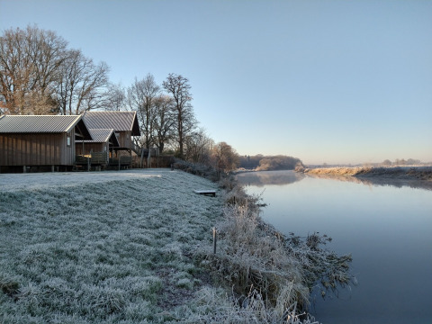 Césped cubierto de escarcha y cabañas de madera junto al río en River Cottage, Camping de Koeksebelt, Países Bajos.