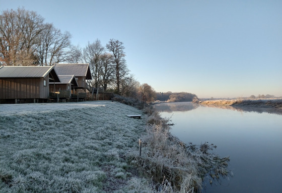 Césped cubierto de escarcha y cabañas de madera junto al río en River Cottage, Camping de Koeksebelt, Países Bajos.