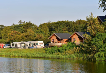 Zicht op River Cottage lodges en caravans bij het meer op Camping de Koeksebelt in Nederland, omgeven door natuur.