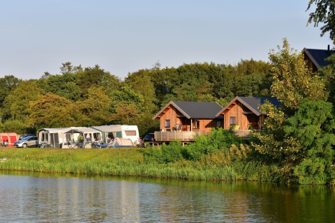 Vista de cabañas River Cottage y caravanas junto al lago en Camping de Koeksebelt, Países Bajos, rodeadas de naturaleza.