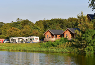 Vista de cabañas River Cottage y caravanas junto al lago en Camping de Koeksebelt, Países Bajos, rodeadas de naturaleza.