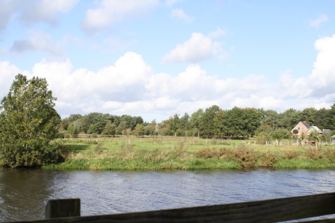 Vista su un fiume tranquillo, campi verdi e una casa al River Cottage, Camping de Koeksebelt, Paesi Bassi.