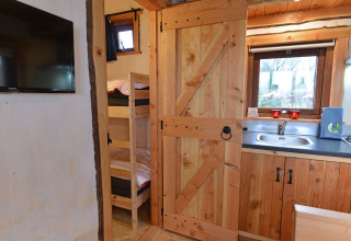 Vista interior de River Cottage en Camping de Koeksebelt, con cocina, literas y televisor en la pared.