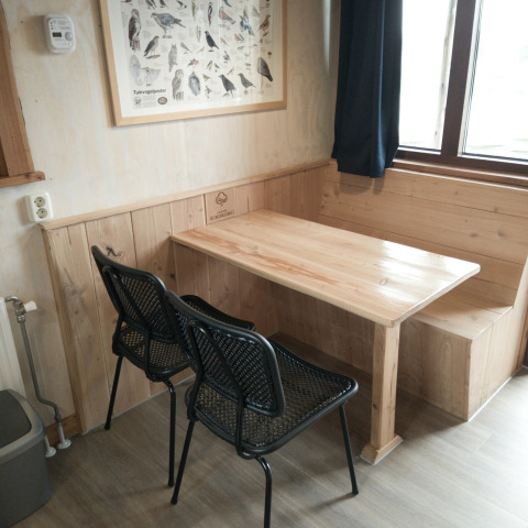 Rincón comedor con mesa de madera, banco, dos sillas negras y póster de aves en River Cottage, Camping de Koeksebelt.
