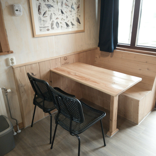 Rincón comedor con mesa de madera, banco, dos sillas negras y póster de aves en River Cottage, Camping de Koeksebelt.