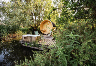 Pod al aire libre con bañera de madera y tumbonas junto al lago, rodeado de vegetación frondosa y árboles.