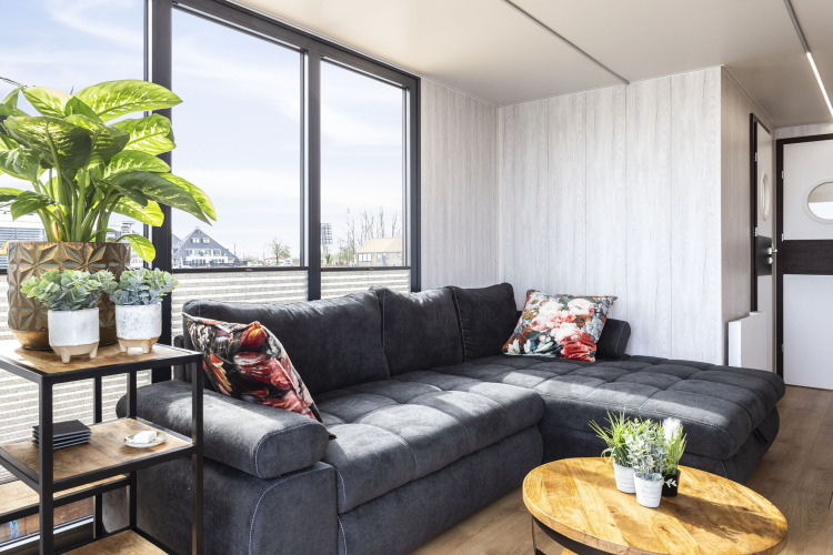 Sala moderna en la casa flotante 'Iere Fügel' con grandes ventanales, sofá gris y plantas, Friesland, Países Bajos.