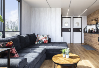 Modern houseboat living room with dark sectional sofa, floral pillows, and kitchenette in Friesland, Netherlands.