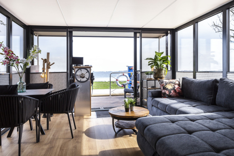 Salón moderno en una casa flotante con ventanales, sofá gris y vistas al lago en Frisia, Países Bajos.