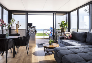 Moderne woonboot met zithoek, grote ramen, grijze zetel en mooi uitzicht op het meer in Friesland.