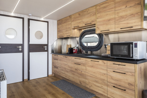 Modernes Hausbootküche mit Holzschränken, rundem Fenster, Mikrowelle und LED-Deckenbeleuchtung.