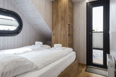 Slaapkamer op woonboot 'Iere Fügel' met twee bedden, houten kast, raam en deur naar terras in Friesland.