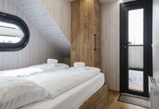 Schlafzimmer auf Hausboot 'Iere Fügel' mit zwei Betten, Holzschrank, Fenster und Tür zur Terrasse in Friesland.