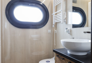 Baño moderno de casa flotante con ventana ovalada, lavabo blanco, muebles de madera y espejo en Friesland.