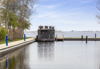 Casa flotante 'Iere Fügel' amarrada en Friesland, Países Bajos, rodeada de agua y paisaje tranquilo.