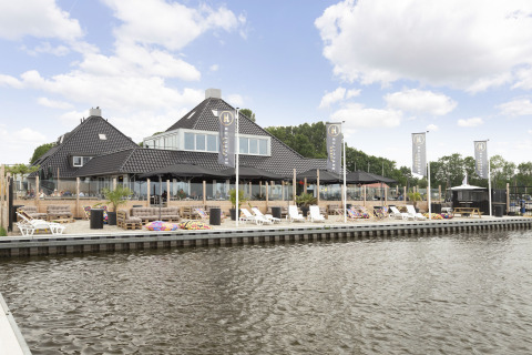 Haus mit schwarzem Dach am Wasser, Liegestühlen und Terrasse bei Houseboat 'Iere Fügel' in Friesland, Niederlande.