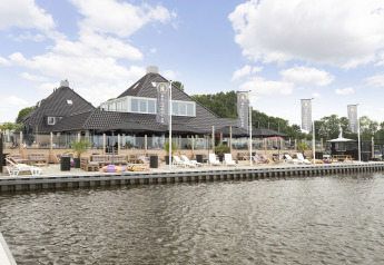 Haus mit schwarzem Dach am Wasser, Liegestühlen und Terrasse bei Houseboat 'Iere Fügel' in Friesland, Niederlande.