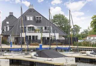 Veleros atracados frente a un moderno houseboat en Friesland, Países Bajos, bajo cielo parcialmente nublado.