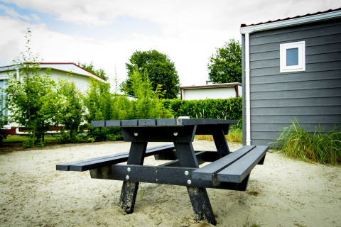 Black picnic bench on sandy ground by Dune Lodge at Holiday Park Duinhoeve, Netherlands, with cabins visible.