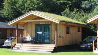 Tiny wooden house with grass roof called Cuckoo's Nest at Camping de Koeksebelt, Netherlands, with cars nearby.