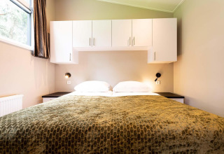 Chambre de lodge avec un grand lit, placards blancs au-dessus, couvertures vertes et lampe murale.