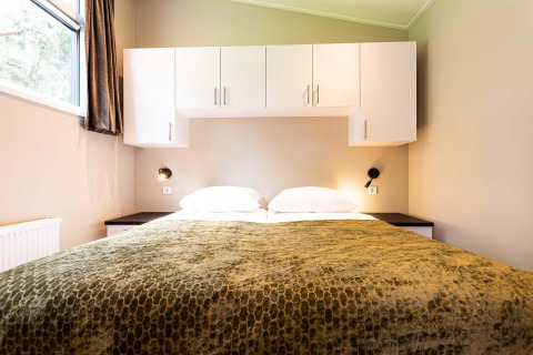 Lodge slaapkamer met tweepersoonsbed, groene deken, witte hangkasten en leeslampjes aan de muur.