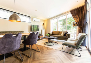 Modern lodge living room with large windows, green couch, dining area, and stylish lighting, nature view.