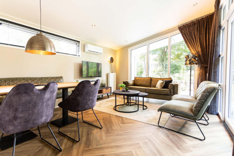Modern lodge living room with large windows, green couch, dining area, and stylish lighting, nature view.