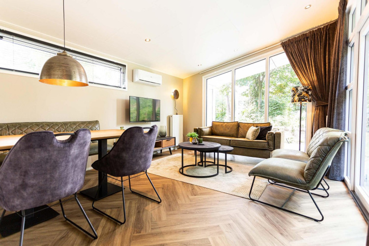 Salón de lodge moderno con grandes ventanales, sofá verde, comedor y elegante iluminación, vista natural.