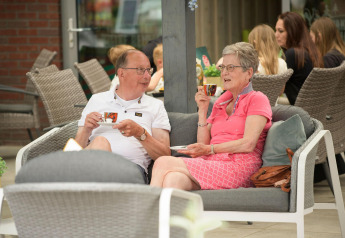 Ældre par nyder kaffe på en terrasse ved glamping-indkvartering, omgivet af andre gæster og komfortable stole.