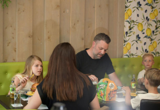 A family of four shares a meal at a table in a glamping accommodation with green seating and wood walls.