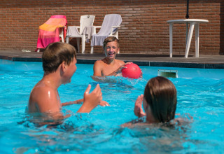Drie kinderen spelen met een bal in het zwembad bij een glampingaccommodatie met stoelen en handdoeken achter hen.