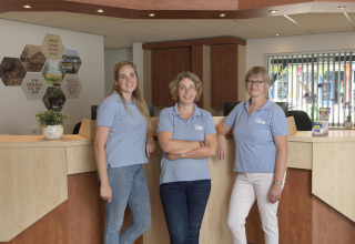 Three female staff members in blue shirts smile at the reception desk of a glamping accommodation.