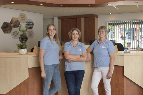 Three female staff members in blue shirts smile at the reception desk of a glamping accommodation.