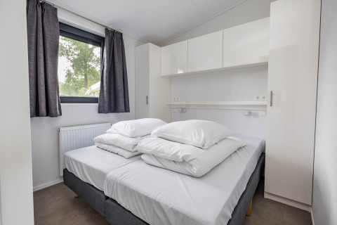 Slaapkamer met dubbel bed, witte kasten en raam in Forest Lodge DeLuxe, Kampeerdorp de Zandstuve.
