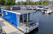 Moderna casa flotante azul Havenlodge N atracada en Marina Parcs Naarden en los Países Bajos con barcos cerca.