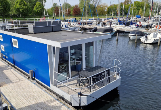 Moderne blauwe woonboot Havenlodge N aangemeerd bij Marina Parcs Naarden in Nederland, met jachten vlakbij.