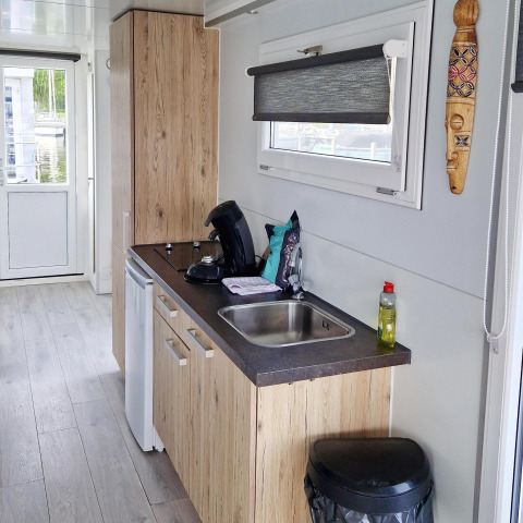 Cocina compacta con fregadero, cafetera y gabinetes de madera en la casa flotante Havenlodge N de Marina Parcs Naarden.