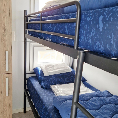 Stapelbed met blauwe lakens op een woonboot, raam achteraan en houten kast links van het bed.