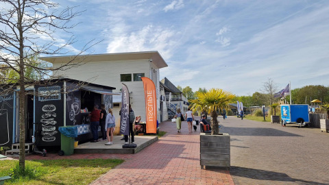 Photo of a glamping accommodation with a food stall, people relaxing, and potted palm trees outside.