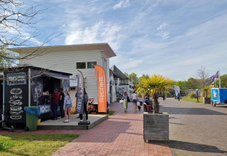 Photo of a glamping accommodation with a food stall, people relaxing, and potted palm trees outside.