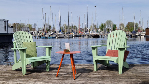 Two green chairs and a table on a wooden dock by the water with a view of a marina on a sunny day.